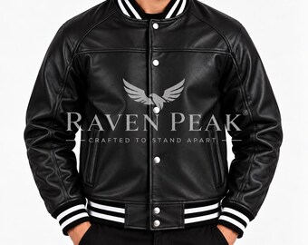 May include: A black varsity-style jacket featuring a smooth finish, silver-toned snap buttons, and a ribbed collar and cuffs with white stripes. The chest displays a grey eagle graphic above the text RAVEN PEAK CRAFTED TO STAND APART.