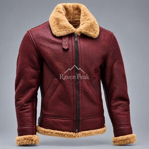 Puede incluir: Una chaqueta de color burdeos oscuro con cuello, puños y dobladillo de borreguillo beige grueso. La chaqueta tiene una cremallera central negra, dos bolsillos delanteros inclinados y una pequeña lengüeta en el cuello. El texto Raven Peak está impreso en blanco.