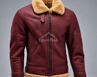 May include: A deep burgundy jacket featuring a thick tan shearling collar, cuffs, and hem. The jacket has a central black zipper, two slanted front pockets, and a small tab at the neck. The text Raven Peak is printed in white on the chest.