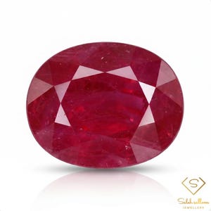 May include: A deep red, oval-cut gemstone with a faceted surface that reflects light. The stone displays a rich, saturated crimson hue throughout its structure. In the bottom right corner, there is a logo with the text Saleh sallom jewellery.