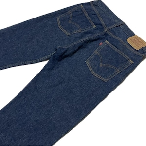 May include: Dark blue denim jeans featuring two back pockets with arcuate stitching, a tan leather-like patch on the waistband, and a small red tab attached to the right pocket. The jeans are constructed with visible orange contrast stitching.