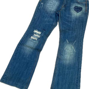 Pode incluir: Calças de ganga em tom azul médio com acabamento desgastado, apresentando cortes horizontais desfiados na perna direita, uma marca desbotada em forma de x na perna esquerda e um emblema azul escuro em forma de coração com pontos brancos.