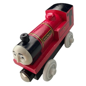 Op de afbeelding: Een rode houten speelgoedtrein met een zwarte voorkant en dak, voorzien van een wit gezicht met zwarte wenkbrauwen en ogen. Op de zijkant van de rode ketel staat de naam RHENEAS in gele letters en het nummer 2 op het achterpaneel.