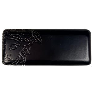 May include: A rectangular black case with rounded corners, featuring a raised, embossed Medusa head design on the left side. The surface has a smooth, matte finish, and the overall construction is solid with a clean, minimalist aesthetic.