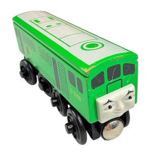 Könnte beinhalten: Eine grüne Spielzeugeisenbahn-Lokomotive aus Holz mit schwarzen Rädern und einem weißen Gesicht mit aufgemalten Augenbrauen und Augen. Die Seite des Zuges ist mit dem Text D 5702 in Gelb markiert. Das Dach des Zuges ist weiß mit grünen Details.