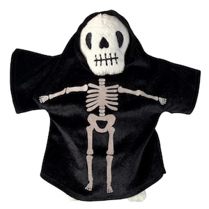 May include: A plush toy featuring a white skull head with black embroidered eyes, nose, and mouth, wearing a black hooded robe. The front of the robe displays a light gray printed skeleton rib cage, arm, and leg bones across the chest and torso.