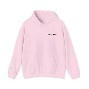 Op de afbeelding: Een lichtroze hoodie met een capuchon met trekkoord en een kangoeroezak aan de voorkant. Het woord "OFFLINE" is in het zwart op de borst gedrukt. De manchet heeft kleine tekst. De hoodie is gemaakt van een zacht materiaal.