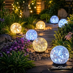 Pode incluir: Cena de jardim com várias luzes de globo de vidro iluminadas em tons de branco quente e azul frio. As luzes estão dispostas ao longo de um caminho de pedra, rodeadas por vegetação e flores roxas, criando uma exibição de iluminação exterior decorativa.