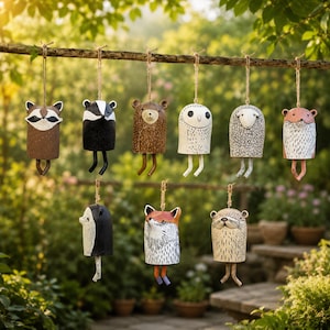 May include: A selection of charming animal-shaped hanging decorations. Each ornament is made of metal and depicts a different animal, such as a raccoon, badger, bear, owl, sheep, and fox. They are suspended from a branch with twine.