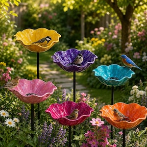May include: A collection of six colorful, flower-shaped bird feeders in a garden setting. The feeders are in shades of yellow, purple, teal, pink, magenta, and orange. Each feeder has a black metal stand and is occupied by a small bird. The garden is filled with flowers.