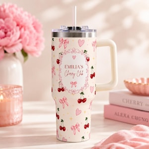 Könnte beinhalten: Ein cremefarbener Edelstahlbecher mit Henkel und Strohhalm, verziert mit einem Muster aus roten Kirschen, rosa Schleifen und kleinen Herzen. In der Mitte befindet sich ein dekorativer Rahmen mit dem Text EMILIA'S Cherry Club in roter Schrift.