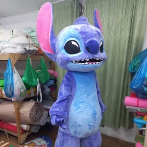 Peut inclure: Un costume en peluche int&eacute;gral d'un personnage de dessin anim&eacute;, principalement dans les tons de bleu et de violet. Le costume pr&eacute;sente de grandes oreilles avec l'int&eacute;rieur rose, un large sourire avec des dents blanches et de grands yeux noirs. Le ventre du personnage est d'un bleu plus clair.