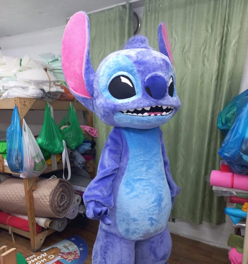 Peut inclure: Un costume en peluche int&eacute;gral d'un personnage de dessin anim&eacute;, principalement dans les tons de bleu et de violet. Le costume pr&eacute;sente de grandes oreilles avec l'int&eacute;rieur rose, un large sourire avec des dents blanches et de grands yeux noirs. Le ventre du personnage est d'un bleu plus clair.
