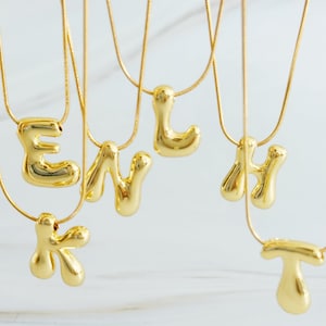 Può includere: Una collezione di collane color oro con ciondoli a forma di lettera dallo stile liquido e gonfio. Ogni collana è composta da una sottile catena a serpente flessibile che sostiene un singolo ciondolo iniziale liscio, riflettente e organico.