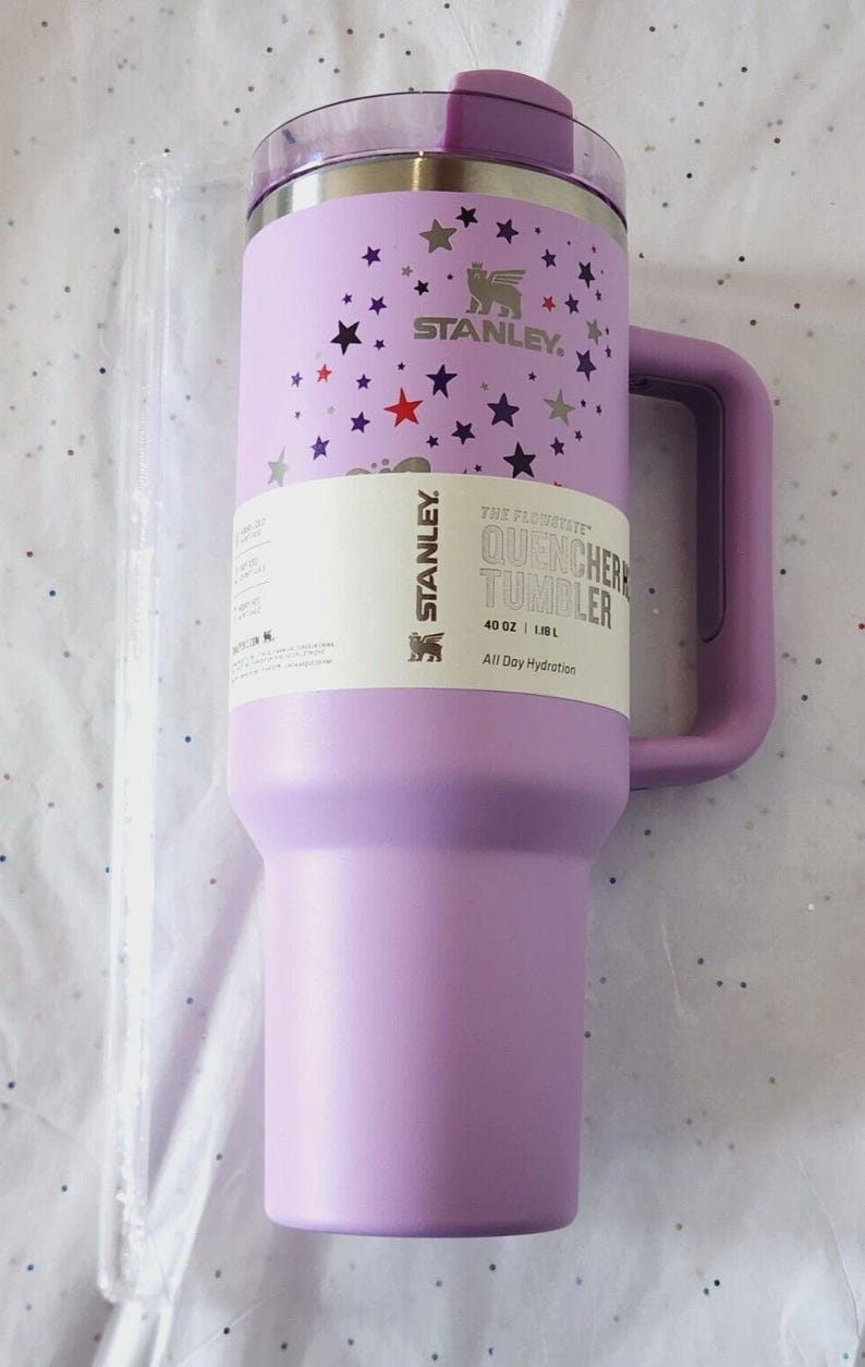 Puede incluir: Un vaso Stanley Quencher lavanda con asa, decorado con estrellas. El vaso tiene una capacidad de 1,18 L y presenta el logotipo de Stanley y el texto "All Day Hydration".