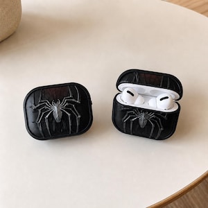 May include: Black protective case for wireless earbuds, with a raised silver spider design. The case is shown closed and open, revealing white earbuds inside. The case is on a light-coloured surface.