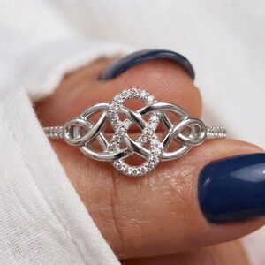 May include: A silver-toned ring featuring an intricate, interwoven knot design. The central portion of the knot is embellished with small, clear, sparkling stones, while the band itself is thin and polished, creating a continuous, detailed pattern.