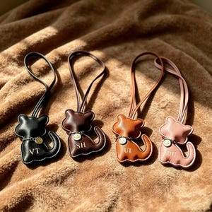 Può includere: Quattro ciondoli per borsa a forma di gatto in materiale liscio, ciascuno con un cinturino ad anello abbinato. I ciondoli sono neri, marrone scuro, marrone chiaro e rosa pallido, con una catenella argentata e le iniziali VT, SH, UI e DE.