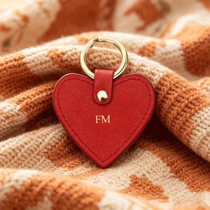 May include: A red heart-shaped keychain with a textured surface and gold-tone hardware. The keychain features the letters FM embossed in gold in the center and includes a circular gold-tone ring for attachment to keys or other items.