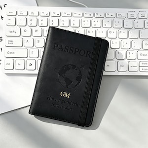 May include: A black passport holder featuring an elastic closure strap. The cover is embossed with the word PASSPORT, a globe graphic with a small airplane, the letters GM in gold, and the text Have a good trip travel wallet.