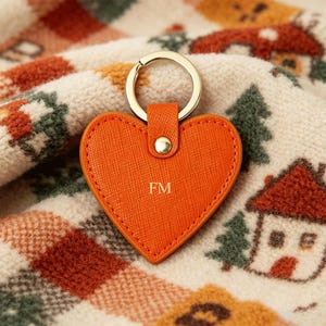 May include: A heart-shaped orange keychain with a textured surface and gold-tone metal ring. The keychain features a small gold-tone rivet at the top and the letters FM embossed in gold in the center of the heart-shaped leather-like material.