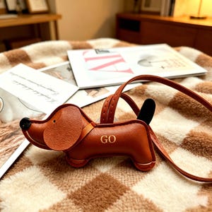 May include: A brown dog-shaped bag charm with a black nose and tail, featuring the letters GO embossed in gold on its side. The charm includes a thin matching brown leather strap for attachment, showcasing detailed stitching along the edges.