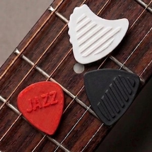 Könnte beinhalten: Drei Gitarrenplektren auf einem Griffbrett. Ein rotes Plektrum mit weißer Aufschrift "JAZZ", ein weißes Plektrum mit einzigartiger Form und ein schwarzes Plektrum mit strukturierter Oberfläche. Das Griffbrett ist aus dunklem Holz.