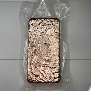 May include: A rectangular, copper-coloured bar with a textured surface, sealed in a clear plastic package. The bar's surface has a wrinkled appearance, with variations in tone and highlights.