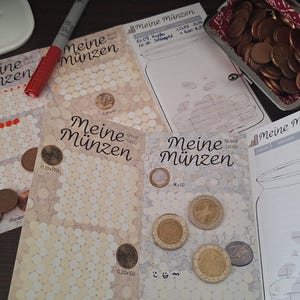 Könnte beinhalten: Mehrere Papierbögen mit der Aufschrift Meine Münzen mit aufgedruckten Münzumrissen zur Sparverfolgung, begleitet von einem mit Kupfermünzen gefüllten Metall-Geldbeutel, losen Münzen auf der Oberfläche sowie einem roten und einem grauen Stift.