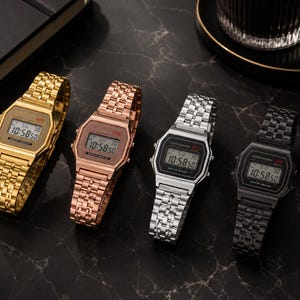 Puede incluir: Cuatro relojes de pulsera digitales en oro, oro rosa, plata y negro. Cada reloj tiene una esfera rectangular que muestra la hora, con una correa de metal a juego. Los relojes se muestran sobre una superficie oscura y texturizada.