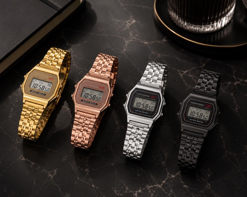 Puede incluir: Cuatro relojes de pulsera digitales en oro, oro rosa, plata y negro. Cada reloj tiene una esfera rectangular que muestra la hora, con una correa de metal a juego. Los relojes se muestran sobre una superficie oscura y texturizada.
