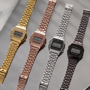 Puede incluir: Cuatro relojes digitales con correas met&aacute;licas en dorado, oro rosa, plateado y gris oscuro. Cada reloj muestra la hora 10:58:50 en su pantalla digital. Los relojes est&aacute;n dispuestos diagonalmente sobre una superficie reflectante.