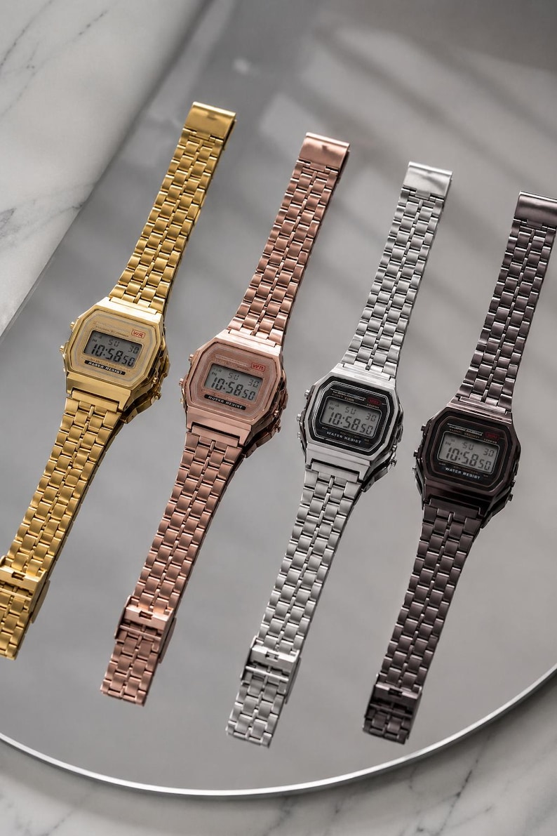 Puede incluir: Cuatro relojes digitales con correas met&aacute;licas en dorado, oro rosa, plateado y gris oscuro. Cada reloj muestra la hora 10:58:50 en su pantalla digital. Los relojes est&aacute;n dispuestos diagonalmente sobre una superficie reflectante.