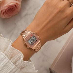 Puede incluir: Reloj digital de oro rosa con una correa a juego. La esfera del reloj muestra la hora y la fecha. El reloj se lleva en la mu&ntilde;eca. El fondo incluye una superficie de m&aacute;rmol y una rosa rosa.