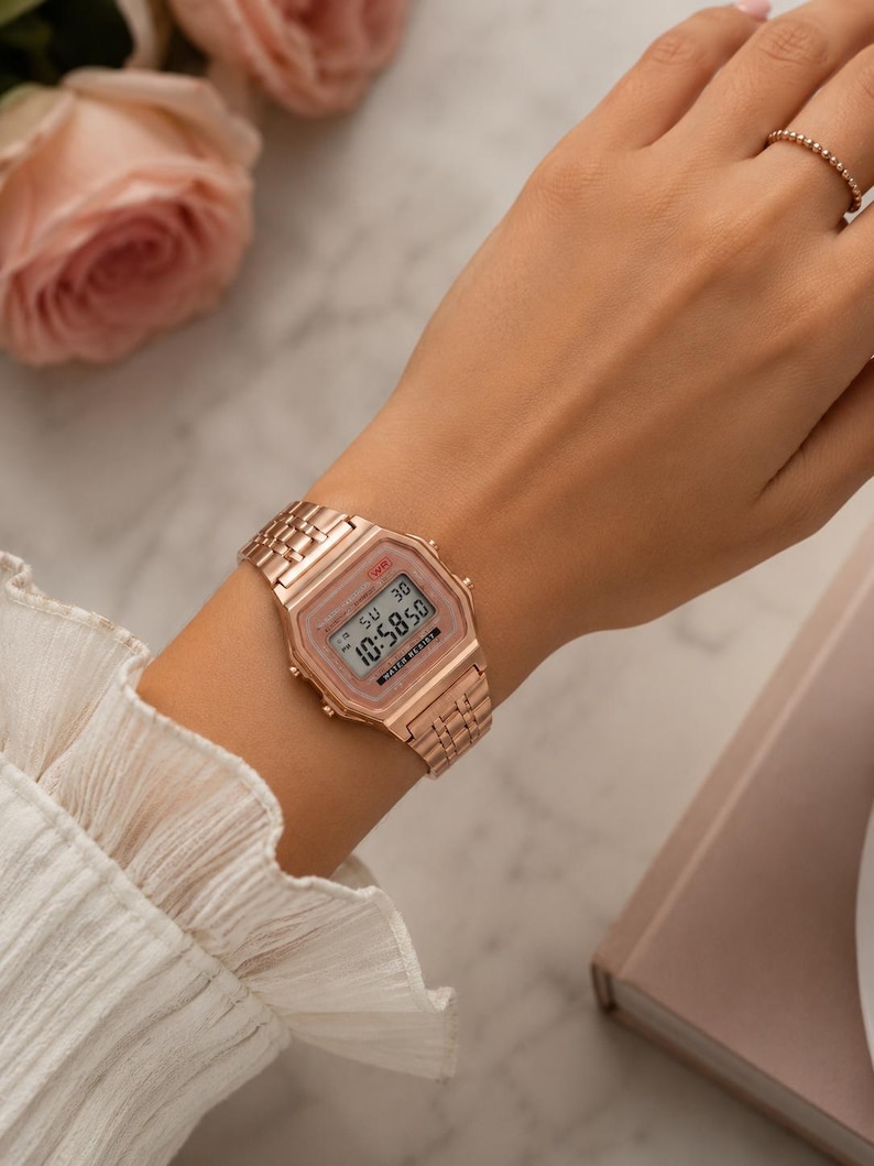 Puede incluir: Reloj digital de oro rosa con una correa a juego. La esfera del reloj muestra la hora y la fecha. El reloj se lleva en la mu&ntilde;eca. El fondo incluye una superficie de m&aacute;rmol y una rosa rosa.