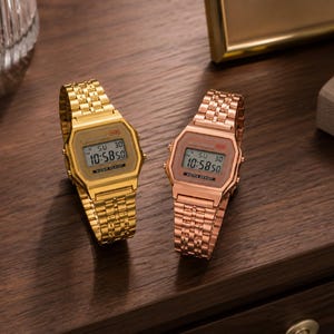 Puede incluir: Dos relojes digitales con correas de metal dorado y oro rosa se exhiben sobre una superficie de madera oscura. Los relojes muestran la hora 10:58:50. Las correas tienen un dise&ntilde;o de eslabones.