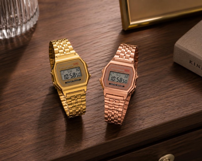Puede incluir: Dos relojes digitales con correas de metal dorado y oro rosa se exhiben sobre una superficie de madera oscura. Los relojes muestran la hora 10:58:50. Las correas tienen un dise&ntilde;o de eslabones.
