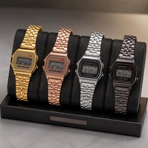 Puede incluir: Cuatro relojes digitales en oro, oro rosa, plata y negro, expuestos en un soporte negro. Cada reloj muestra la hora 10:58:50. Los relojes tienen una esfera rectangular y una correa de metal.
