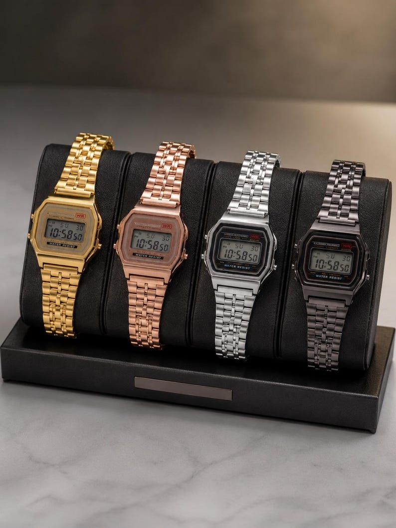 Puede incluir: Cuatro relojes digitales en oro, oro rosa, plata y negro, expuestos en un soporte negro. Cada reloj muestra la hora 10:58:50. Los relojes tienen una esfera rectangular y una correa de metal.