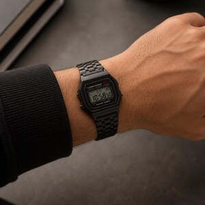 Puede incluir: Un reloj de pulsera digital negro con una correa de metal negro a juego se muestra en una mu&ntilde;eca. La esfera del reloj muestra la hora como 10:58:50. El reloj est&aacute; etiquetado como "WATER RESIST".