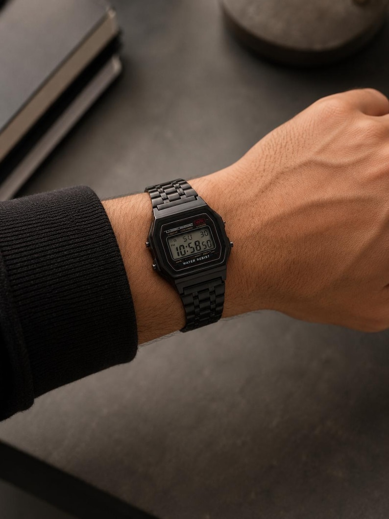 Puede incluir: Un reloj de pulsera digital negro con una correa de metal negro a juego se muestra en una mu&ntilde;eca. La esfera del reloj muestra la hora como 10:58:50. El reloj est&aacute; etiquetado como "WATER RESIST".