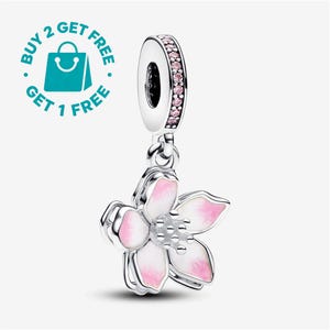 Peut inclure: Une breloque pendante en argent représentant une fleur à cinq pétales avec des détails en émail blanc et rose et un centre en perles d'argent. La bélière est incrustée de petites pierres roses. L'image contient le texte BUY 2 GET 1 FREE.