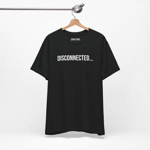 Peut inclure: T-shirt noir avec l'inscription blanche "DISCONNECTED..." centrée sur la poitrine. Le t-shirt est suspendu à un cintre en bois. La marque "ZERO STATE" est imprimée au-dessus du texte.