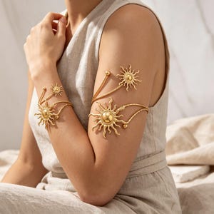 Può includere: Bracciali dorati con disegni a forma di sole. I gioielli includono un bracciale per la parte superiore del braccio e un braccialetto per il polso, collegati da delicate catene. I motivi solari hanno raggi dettagliati e un globo centrale.