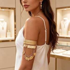 Puede incluir: Brazalete dorado con colgantes de monedas. El brazalete presenta múltiples bandas texturizadas y se lleva en la parte superior del brazo. También se ven pendientes y un collar dorados a juego.