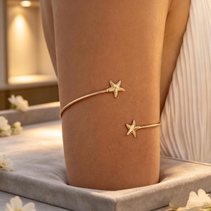 May include: Gold-coloured arm cuff with two star details. The cuff curves around the arm, with a star at each end. The jewellery is displayed on a neutral background, highlighting the design's simplicity and elegance.