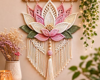 May include: A macrame wall hanging in the shape of a lotus flower. The flower features pink, cream, and green colors, with a wooden dowel at the top for hanging. A tassel hangs from the bottom. The wall hanging is a decorative piece.