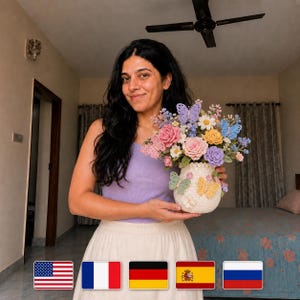May include: A crocheted vase filled with colorful crocheted flowers and butterflies. The flowers include pink roses, purple and yellow blooms, and blue and white accents. The vase is cream-colored with butterfly details.