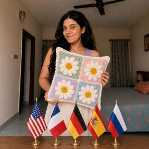 May include: A square crochet pillow featuring four daisy flower designs. Each flower has a yellow center and white petals, set against pastel squares of green, pink, purple, and blue. The pillow has a cream-colored border.