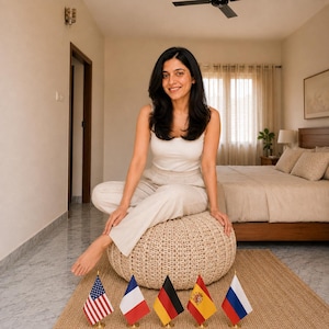May include: A woman sits on a woven ottoman in a bedroom, smiling at the camera. She wears a white tank top and light-colored pants. Small flags of the United States, France, Germany, Spain, and Russia are displayed on a jute rug.
